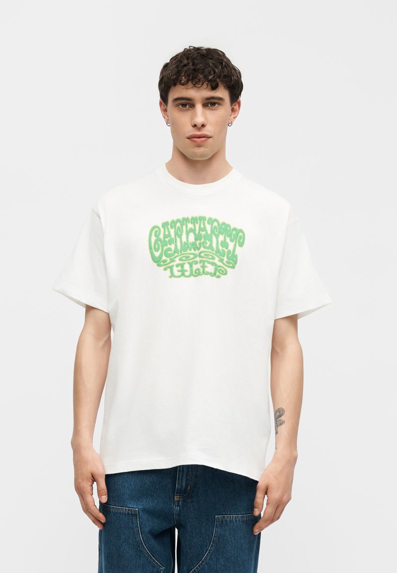 Giovane uomo con capelli corti ricci e scuri, che indossa una maglietta bianca oversize con la scritta verde "GANTHAPPY LIGHT" e jeans blu, in piedi su sfondo bianco.