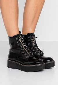 Bottes de plateforme en cuir verni noir brillant avec des semelles épaisses, des zippers argentés et des lacets noirs portées sur des jambes nues contre un fond clair.