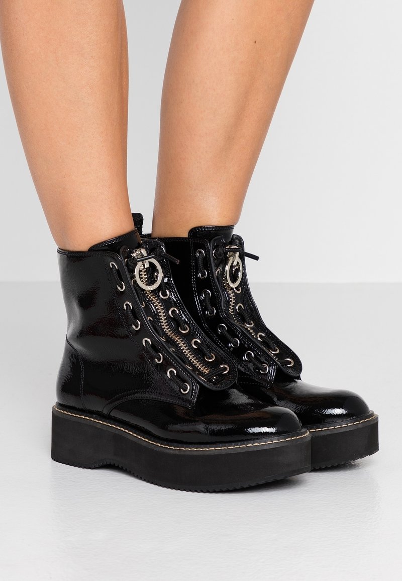 Bottes de plateforme en cuir verni noir brillant avec des semelles épaisses, des zippers argentés et des lacets noirs portées sur des jambes nues contre un fond clair.