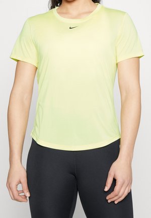 Frau trägt ein hellgelbes kurzärmeliges Sporttop mit kleinem schwarzem Nike-Logo zentriert nahe dem Ausschnitt, kombiniert mit schwarzen Leggings.