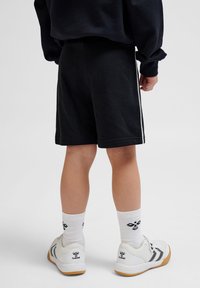 Svarta svettshorts med vita sidosträck, tillsammans med vita träningsskor och mönstrade strumpor, sedd bakifrån.