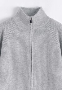 Pull en tricot gris avec un col haut, doté d'une fermeture éclair sur toute la longueur et d'une texture côtelée. Design simple et fonctionnel sans motifs.