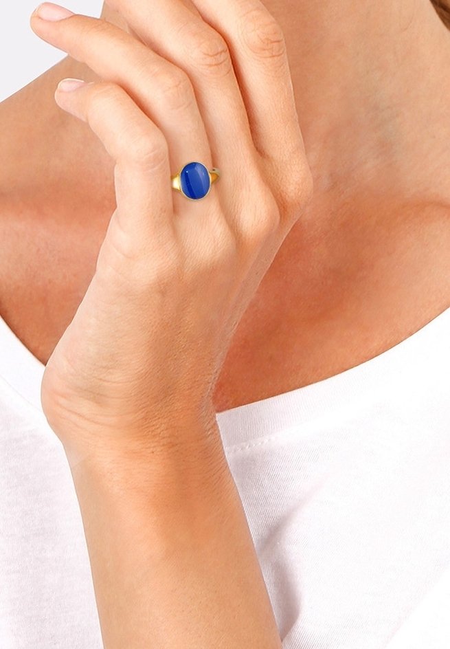 Main avec une bague en or ornée d'une pierre précieuse bleue ovale, tenue près du cou, portant un haut blanc.
