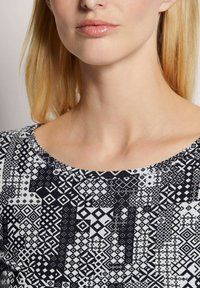 Schwarz-weißer bedruckter Oberteil mit geometrischen Formen, einem runden Ausschnitt und einer glatten Stoffstruktur, die am Schlüsselbein sichtbar ist.