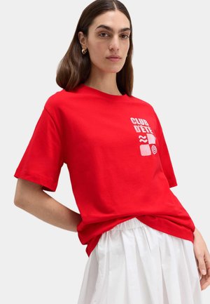 Femme aux longs cheveux bruns portant des boucles d'oreilles en cercle dorées, un T-shirt rouge avec du texte blanc et des graphismes, ainsi qu'une jupe plissée blanche.