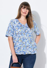 Blusa floral de manga corta en azul, amarillo y blanco con escote en V. Tejido ligero, corte holgado y ligero drapeado. Llevada con jeans oscuros y un bolso marrón.