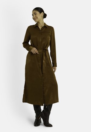Robe chemise marron à manches longues avec col à revers, devant boutonné et taille ceinturée. Les fentes latérales améliorent la mobilité. Accompagnée de bottes noires montantes.