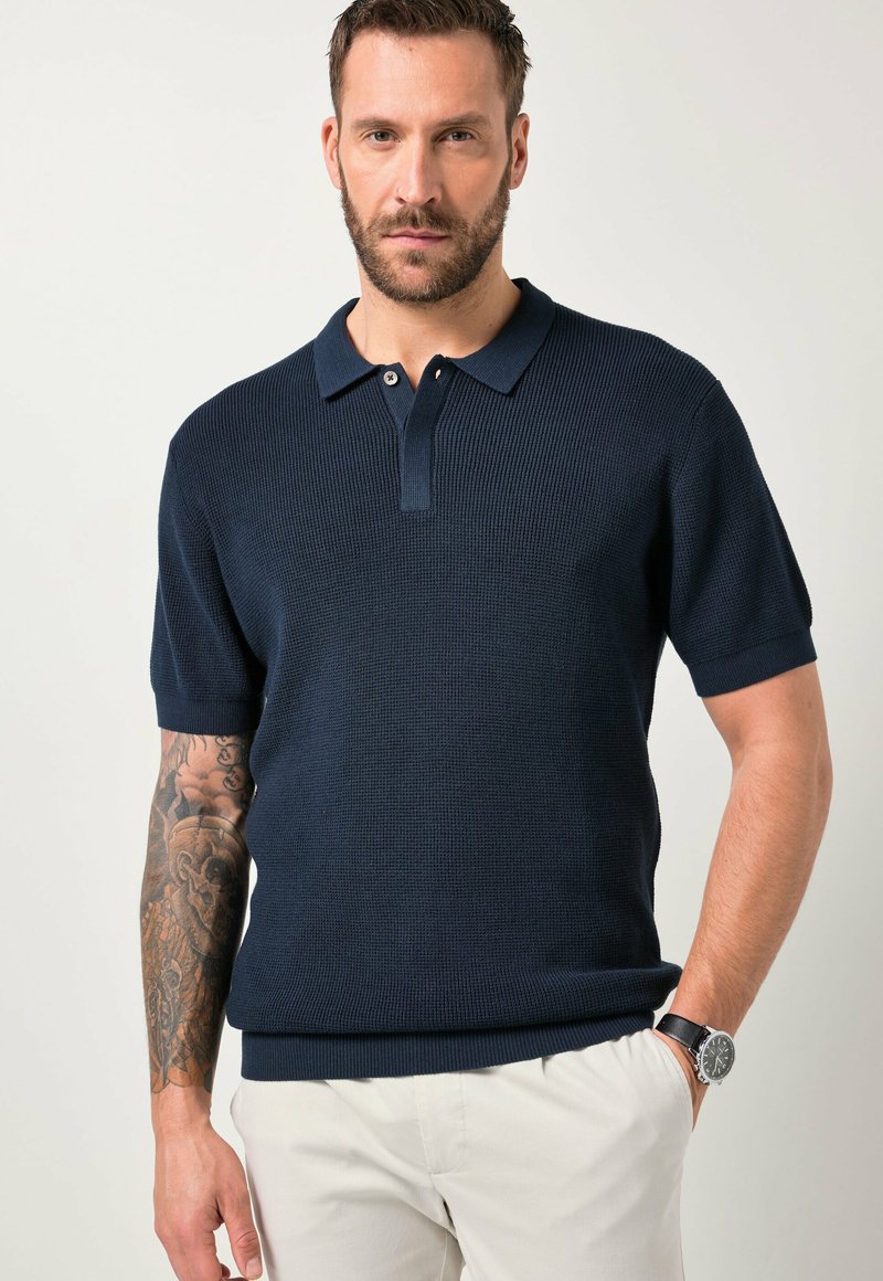 Mand med tatoveret arm iført navy kortærmet tekstureret polo skjorte og hvide bukser, stående med den ene hånd i lommen mod en ensfarvet baggrund.