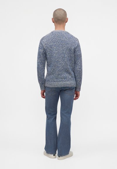 Homme à tête rasée portant un pull en maille bleu chiné, un jean évasé bleu et des baskets blanches, debout de dos sur un fond blanc.
