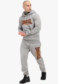 Sivá mikina s kapucňou a tepláky so značkovým logom "LONSDALE LONDON" v oranžovej farbe. Mäkká tkanina, nastaviteľná kapucňa, priliehajúce manžety a elastický pás.