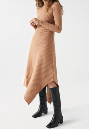 Vestito a maniche lunghe in maglia beige con orlo asimmetrico, abbinato a stivali alti neri in pelle con tacco quadrato.