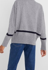 Pull gris en maille ample, avec une bande horizontale bleu marine à l'ourlet et aux manches, associé à un jean en denim bleu.