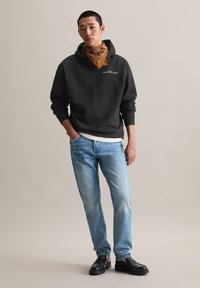 Zwarte hoodie met logo, blauwe spijkerbroek en een bruine bandana. De outfit bestaat uit zwarte schoenen en heeft ontspannen denim en zachte materiaalkleuren.