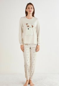 Beige pyjamastel met een lange mouwen shirt waarop een puppy applicatie is gestikt en bijpassende broek met kleine zwarte en gouden hartpatronen.