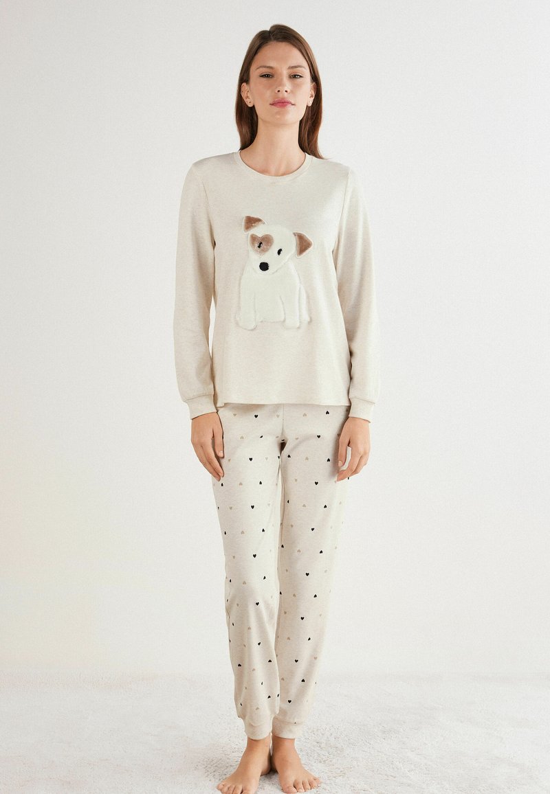 Beige pyjamastel met een lange mouwen shirt waarop een puppy applicatie is gestikt en bijpassende broek met kleine zwarte en gouden hartpatronen.