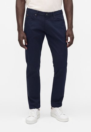 Mand iført mørkeblå slim-fit bukser og hvide sneakers, stående mod en ensfarvet hvid baggrund.