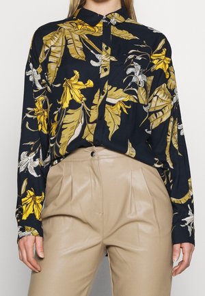 Czarna koszula na guziki z żółto-białym kwiatowym nadrukiem, noszona wpuszczona w beżowe skórzane spodnie z wysokim stanem.