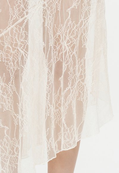 Tissu en dentelle blanche transparente avec broderie florale drapé sur une jambe, montrant une texture délicate et des motifs complexes.