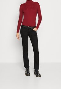 Pull à col roulé côtelé rouge, jean noir coupe droite, bottines en cuir. La tenue est complétée par une fine ceinture noire et des détails minimalistes.