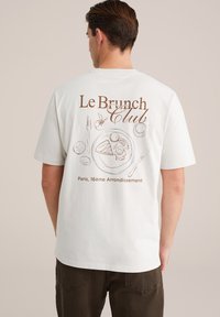Balta kokvilnas t-kreklu ar brūnu grafiku uz aizmugures, kurā attēlota šķīvis ar ēdienu, galda piederumi un teksts "Le Brunch Club". Vienkāršs, ikdienišķs dizains.