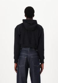 Beskarpet svart hettegenser med snor, ribbestrikkede mansjetter og detaljer i sømmen på baksiden, sammen med høyhælte mørke denimjeans.