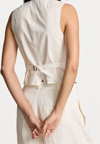 Žena oblečená v tílku světlé barvy se zapínáním na přezku a ladících kalhotách s vysokým pasem, s rukama zkříženýma za zády.