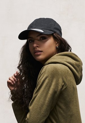 Czarna czapka z zakrzywionym daszkiem, noszona z zieloną polarową bluzą. Tkanina wygląda na miękką; model ma wyraźne rysy twarzy i luźne, kręcone włosy.