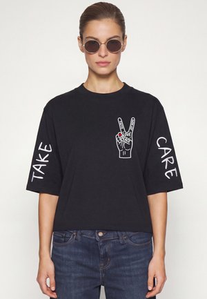Vrouw die ronde zonnebril en een zwart T-shirt draagt met "Take Care" op de mouwen en een vredesteken met woorden op de borst.