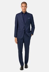 Traje azul marino a medida con un sutil patrón de rayas, chaqueta de un solo pecho, dos botones, solapas de muesca y pantalones a juego, combinado con una camisa oscura.