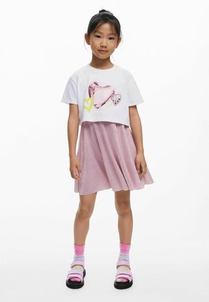 Junges Mädchen steht und trägt ein weißes kurz geschnittenes T-Shirt mit Herzen, einen rosa plissierten Rock, pinke verlaufende Socken und rosa Sandalen.