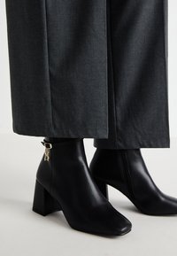 Bottines en cuir noir avec un bout carré et un talon bloc. Présence d'un détail de logo doré sur la sangle. Associées à un pantalon large gris.