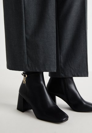 Zwarte leren enkelboots met een vierkante neus en blokhak. Heeft een gouden logo-detail op de band. Gecombineerd met grijze wijde broek.