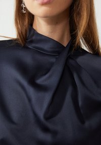 Blouse en satin bleu marine avec un col montant, un détail de tissu torsadé au niveau du col et une texture lisse et douce.