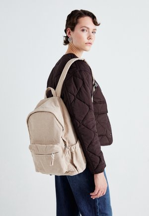 Femme aux cheveux courts portant une veste matelassée sombre et un jean bleu, portant un sac à dos en velours côtelé beige sur une épaule, sur un fond uni.