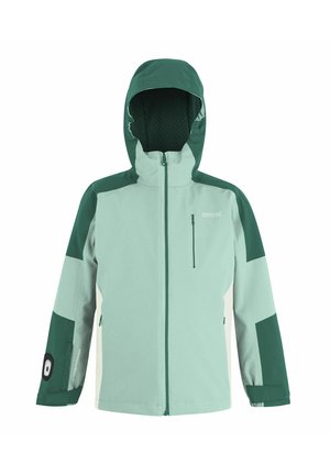 Młoda dziewczyna z długimi blond włosami, ubrana w turkusowo-zieloną kurtkę, czarne spodnie i czarne trampki, stojąca na jednolitym białym tle.