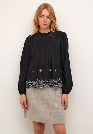Blusa nera a maniche lunghe con collo arricciato e delicata ricamatura bianca all'orlo, abbinata a una gonna leggera e strutturata.