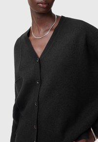 Svart kofta i mjukt material, med V-ringning, knäppning och ribbade ärmslut. Slät textur med minimal detaljering.