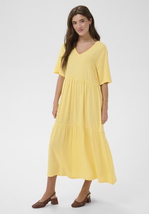 Naine seisab, kandes kollast V-kaelusega kihilist midi kleiti lühikeste varrukatega ja pruune plokksäärega punutud slingback-kingi.