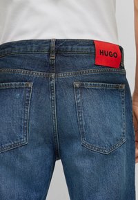 Bakvy av blå denimjeans med orange sömmar och två bakfickor, med en röd "HUGO"-logg på midjan.