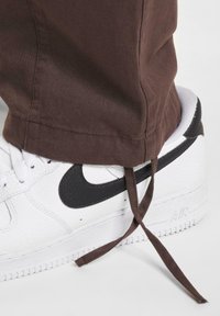 Zapatilla Nike Air Force 1 blanca con swoosh negro, cuero texturizado y cordones marrones. El dobladillo de unos pantalones marrones reposa sobre el zapato.