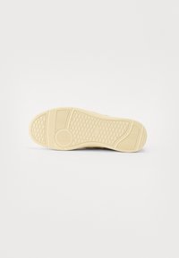 Sneakersåle med en teksturert, beige gummioverflate. Den har et mønstrete mønster for grep og runde former ved hæl- og tåseksjoner.