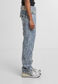 Jeans de mezclilla azul claro con acabado desgastado, que presentan un corte recto, costuras en contraste y bolsillos traseros bordados. Zapatillas plateadas.