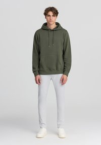 Felpa verde oliva con tasca a marsupio, cordini regolabili, polsini a coste, abbinata a jeans grigi chiari e sneakers bianche.