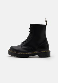 Bottines en cuir noir avec un bout arrondi, un laçage à l'avant, des surpiqûres jaunes et une semelle en caoutchouc texturée. Équipées d'une languette à l'arrière.
