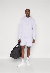 Felpa lilla con cappuccio, tasca frontale e cordini, pantaloni corti coordinati, calze bianche e sneakers, mentre tiene un borsone nero.