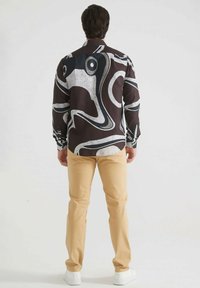 Chemise pour homme en tissu marron foncé avec des motifs abstraits blancs et noirs. Associée à un pantalon beige clair et des baskets blanches.