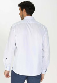 Chemise blanche à manches longues avec col boutonné, texture en coton lisse et détail de logo subtil sur le poignet. La vue arrière révèle une coupe structurée.