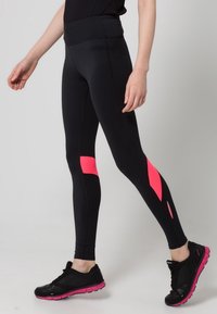 Gambe che indossano leggins neri con accenti geometrici rosa e scarpe da ginnastica nere con suole rosa, in piedi contro uno sfondo semplice.