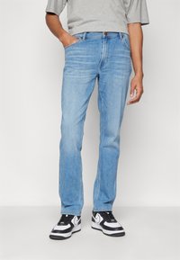 Jeans de denim azul claro con un diseño de pierna recta, cinco bolsillos y cierre de cremallera, combinados con zapatillas negras y blancas.