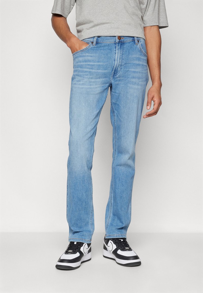 Jeans de denim azul claro con un diseño de pierna recta, cinco bolsillos y cierre de cremallera, combinados con zapatillas negras y blancas.